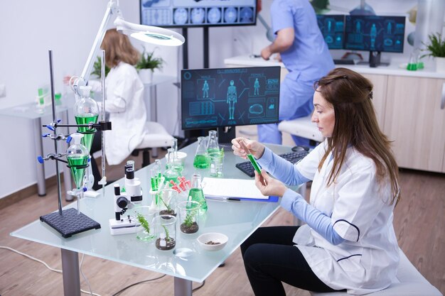 Femme scientifique tenant une solution verte pendant que l'équipe d'écoute travaille en arrière-plan. Jeune biologiste en arrière-plan.