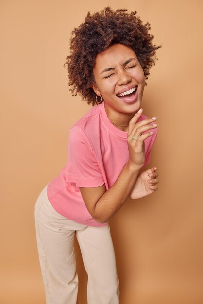 la femme se penche en avant rit sincèrement les yeux fermés à une blague hilarante porte un t-shirt rose décontracté et un pantalon isolé sur beige. Notion d'émotions