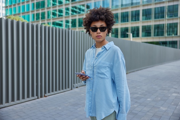 une femme se promène avec un téléphone à l'extérieur porte des lunettes de soleil et une chemise bleue se promène pendant les loisirs dans l'urbanité de la ville utilise des chats d'applications mobiles dans les médias sociaux concentrés loin