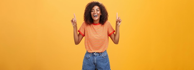 Femme se sentant amusée et divertie Portrait d'une fille afro-américaine élégante et insouciante avec une coiffure afro riant à haute voix joyeusement pointant vers le haut avec les bras levés sur le mur orange