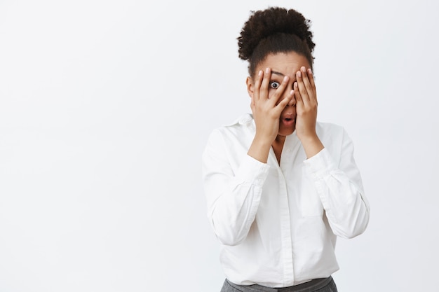 Femme se sentant effrayée mais curieuse, furtivement à travers les doigts avec un œil tout en couvrant la vue avec des paumes, pliant les lèvres de choc et de stupéfaction, debout effrayée sur un mur gris