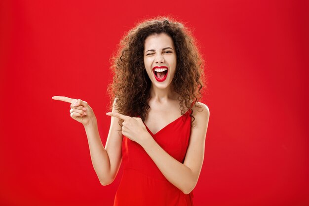 Femme se sentant géniale lors d'une grande fête d'amis pointant vers l'entrée. Élégante femme aux cheveux bouclés et insouciante en robe de soirée rouge s'amusant près de la piscine en souriant joyeusement et en pointant vers la gauche.