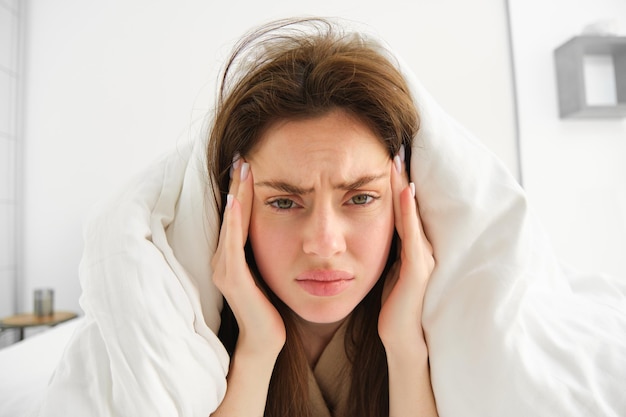 Photo gratuite femme se sentant mal au lit, allongée dans sa chambre sous une couverture et des draps blancs le matin, fronçant les sourcils