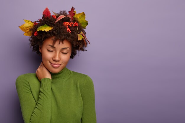 Femme séduisante détendue avec une coiffure afro, touche le cou, a les yeux fermés, a un regard reposant, porte des feuilles dans les cheveux, des vêtements verts