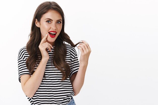 Femme séduisante à la mode et glamour enthousiaste ravie d'une nouvelle coupe de cheveux, porter du rouge à lèvres rouge, toucher curl bouche ouverte impressionné et ravi, satisfait bon produit de soin des cheveux, mur blanc