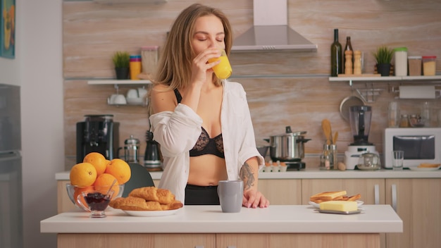 Femme séduisante en sous-vêtements sexy profitant du matin en buvant un verre de jus d'orange frais assis dans la cuisine. Jeune femme avec des tatouages portant de la lingerie noire rafraîchissante dimanche matin