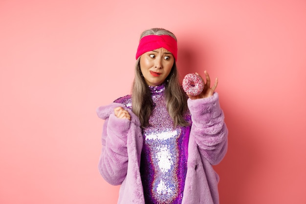 Femme senior asiatique drôle en robe disco scintillante et manteau en fausse fourrure à la tentation de délicieux beignets, voulant manger sucré, debout sur fond rose