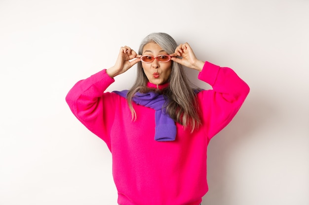 Femme senior asiatique à la mode en lunettes de soleil, lèvres plissées et regardant la caméra surprise, regardant étonnée par l'offre spéciale, debout sur fond blanc.