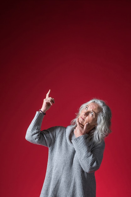 Femme senior contemplée, pointant le doigt vers le haut, debout sur fond rouge