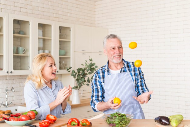 Femme senior excitée applaudissant pendant que son mari jongle avec des citrons dans la cuisine