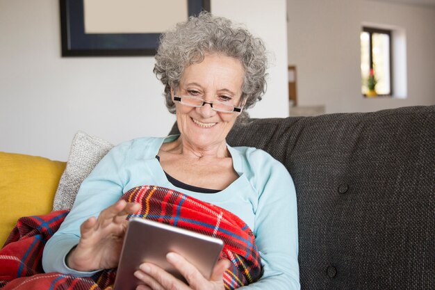 Femme senior joyeuse lire un livre intéressant à l&#39;aide d&#39;une tablette