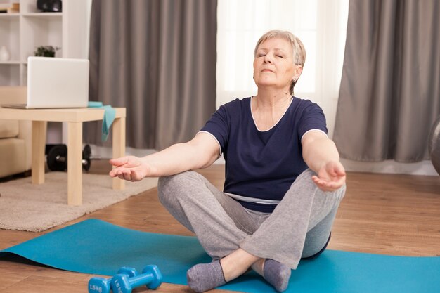 Femme senior paisible avec les yeux fermés faisant du yoga dans le salon