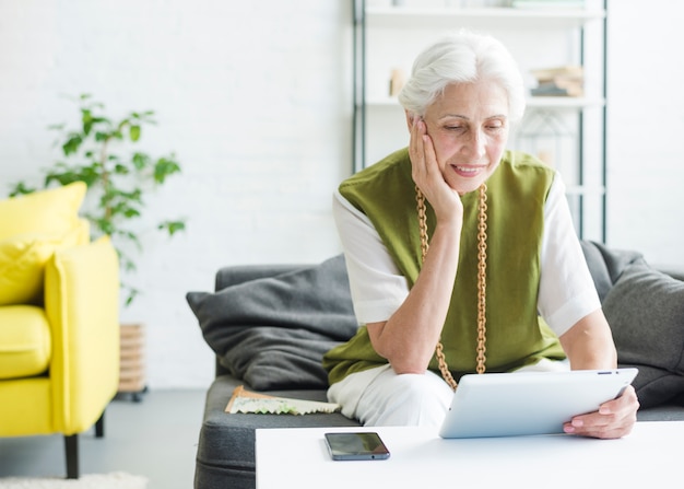 Femme senior souriante assise dans le salon en regardant une tablette numérique