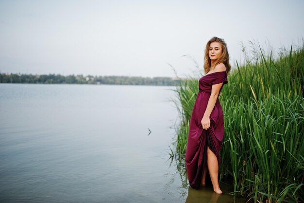 Femme sensuelle blonde en robe marsala rouge debout dans l'eau du lac avec des roseaux