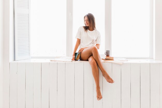 Femme sensuelle avec livre de détente sur le rebord de la fenêtre