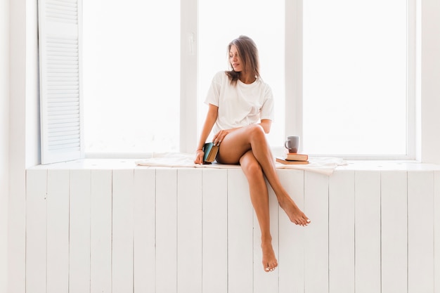 Femme sensuelle avec livre de détente sur le rebord de la fenêtre