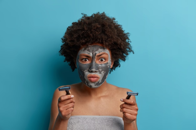 Femme sérieuse avec Afrohair concentré à quelque chose de très attentivement, applique un masque d'argile pour réduire les rides, tient le rasoir pour avoir des procédures hygiéniques après avoir pris une douche isolée sur un mur bleu