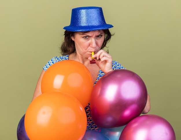 Femme sérieuse d'âge moyen en chapeau de fête avec bouquet de ballons colorés soufflant dans un sifflet avec un visage fronçant les sourcils