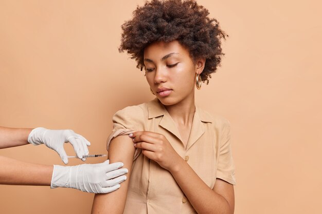 Une femme sérieuse aux cheveux bouclés se fait vacciner dans le bras pour la santé immunitaire isolée sur un mur marron