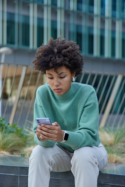 Une femme sérieuse aux cheveux bouclés utilise un smartphone partage une publication de voyage via une application mobile recherche une application Web porte un pull et un pantalon décontractés aime les poses de réseautage mobile à l'extérieur pendant la journée