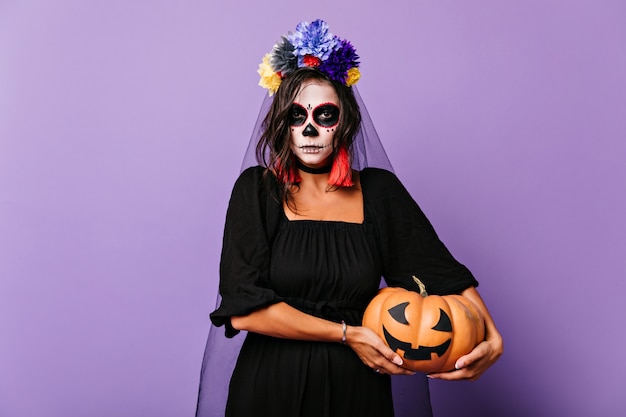 Femme sérieuse aux cheveux bruns en voile tenant la citrouille d'halloween. Plan intérieur d'une fille magnifique en tenue de mariée morte avec un maquillage effrayant.