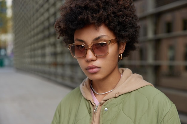 Une femme sérieuse et élégante aux cheveux bouclés porte une veste de lunettes de soleil et un sweat à capuche regarde directement la caméra se promène à l'extérieur pendant la journée se tient sur un arrière-plan flou. Concept de personnes et de style de vie