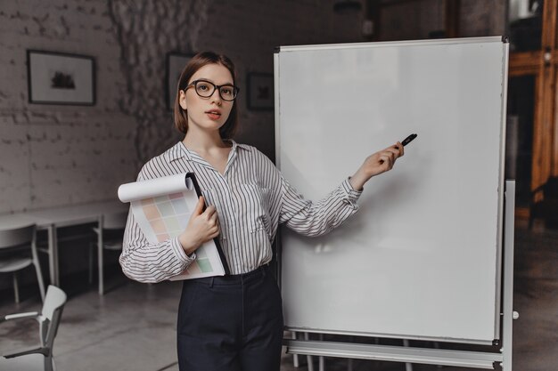 Une femme sérieuse en pantalon noir et chemise rayée parle de l'état des affaires, pointe le marqueur au tableau et tient des rapports.