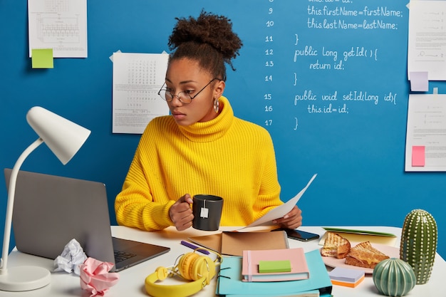 Une femme sérieuse à la peau sombre vérifie les informations contenues dans des papiers et un ordinateur portable, regarde un webinaire de formation sur la programmation informatique