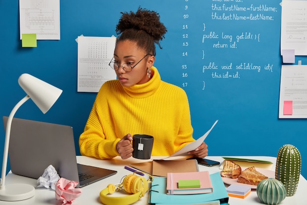 Photo gratuite une femme sérieuse à la peau sombre vérifie les informations contenues dans des papiers et un ordinateur portable, regarde un webinaire de formation sur la programmation informatique