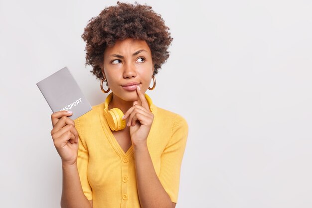 Une femme sérieuse et réfléchie aux cheveux afro considère que les voyages futurs ont l'air pensifs porte un pull jaune décontracté tient le document officiel du passeport pose contre le mur blanc