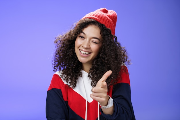Femme sociable et élégante à l'air sympathique avec des cheveux bouclés en bonnet rouge clignotant joyeusement et pointant avec un pistolet à doigt vers la caméra comme compagnon de salutation cool et confiant sur le mur bleu.
