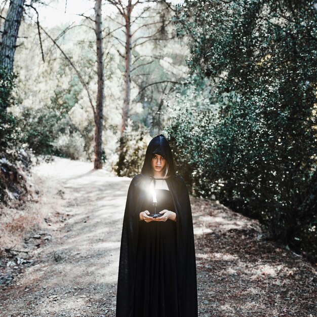 Femme sombre au cap tenant une bougie allumée dans une forêt ensoleillée