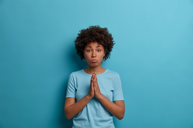 Photo gratuite une femme sombre et bouleversée aux cheveux afro maintient les paumes pressées ensemble pour prier, supplie sur le mur bleu, a besoin de votre aide, demande faveur, porte un t-shirt décontracté, fait une expression de visage innocente