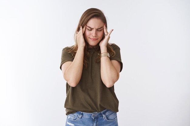 Une femme souffrant d'une énorme migraine sensation douloureuse touchant les tempes ne peut pas retenir la douleur, ferme les yeux en fronçant les sourcils intenses, mal concentrés, les analgésiques forts ne fonctionnent pas, debout malade fond blanc