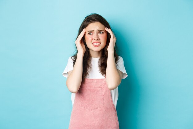 Femme souffrant de migraine douloureuse, touchant la tête et fronçant les sourcils, regardant le coin supérieur gauche, souffrant de maux de tête, debout sur fond bleu