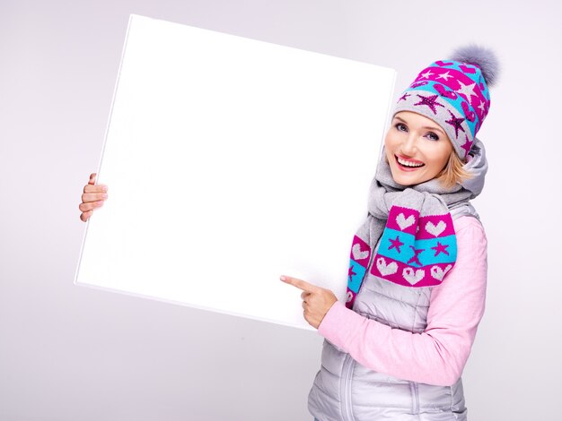 Femme souriante adulte en vêtements d'extérieur chauds tient la pancarte blanche dans les mains et pointe dessus