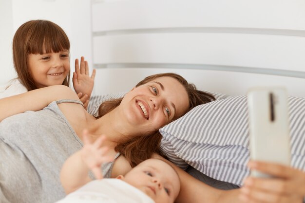Femme souriante allongée dans son lit avec deux filles, diffusant en direct tôt le matin ou ayant un appel vidéo, une fille aînée agitant la main à la caméra, saluant, disant bonjour.