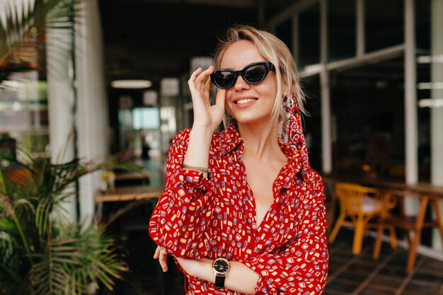 Femme souriante aux cheveux blonds touchant ses lunettes en terrasse d'été