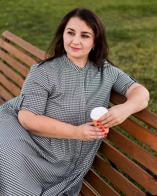 Femme souriante brune tenant une tasse de café alors qu'il était assis sur un banc