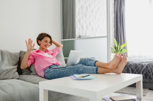 Femme souriante en chemise rose assis détendu sur un canapé à la maison à table travaillant en ligne sur un ordinateur portable à la maison