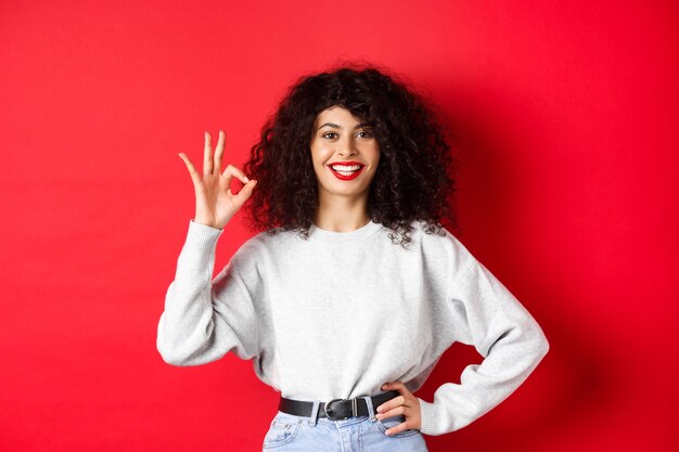 Femme souriante confiante dans des vêtements décontractés, montrant le signe OK pour approuver ou aimer le produit, garantit une bonne qualité, faisant un compliment, fond rouge.