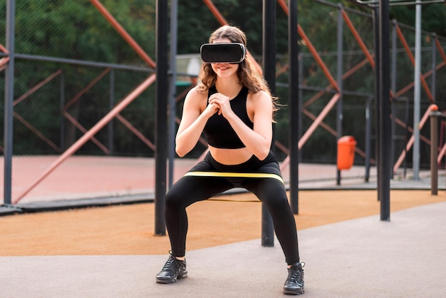 Photo gratuite femme souriante dans un casque vr dans un survêtement faisant des exercices avec un élastique sur un terrain de sport