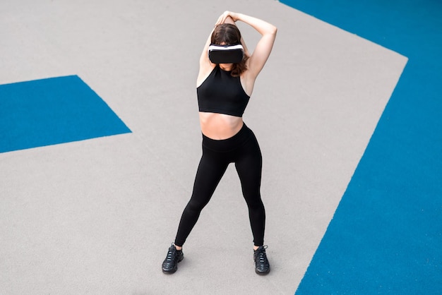 Femme souriante dans un casque VR dans un survêtement faisant des exercices sur un terrain de sport dans un parc