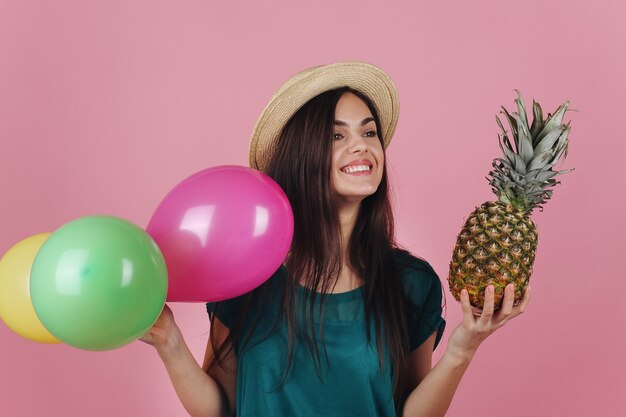 Femme souriante dans un chapeau pose avec des ballons colorés et un ananas