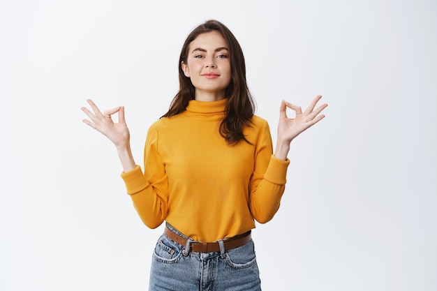 Femme souriante et détendue méditant, ressentant la paix et la patience, respirant l'air librement, debout contre un mur blanc