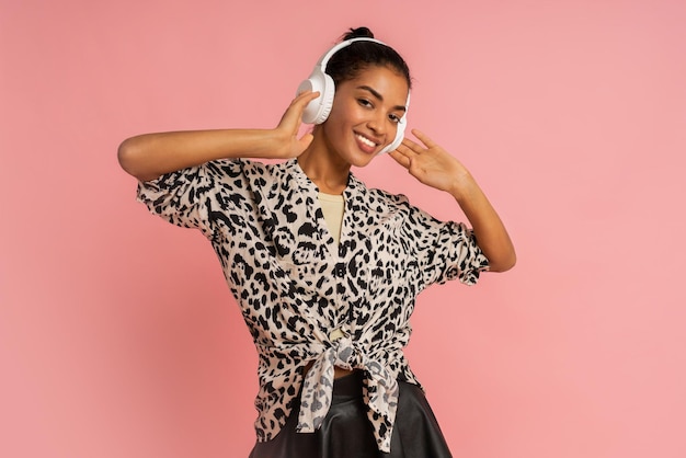 Femme souriante écoutant de la musique par des écouteurs posant sur fond rose portant un chemisier élégant et une jupe en cuir