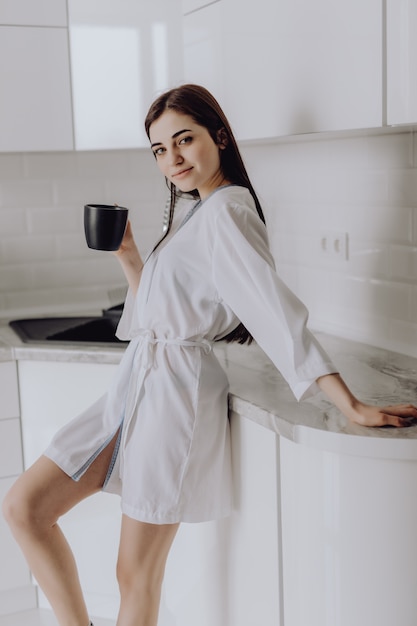 Femme souriante élégante en robe blanche debout devant une cuisine en buvant une tasse de café tôt le matin