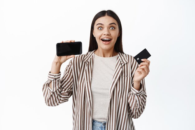 Une femme souriante excitée gagne de l'argent en ligne, montrant une carte de crédit en plastique et un écran vide de smartphone horizontalement, debout dans une tenue élégante contre un mur blanc