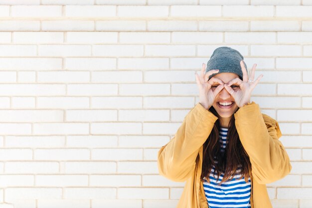 Femme souriante faisant un geste correct comme des jumelles portant une veste et un bonnet tricoté