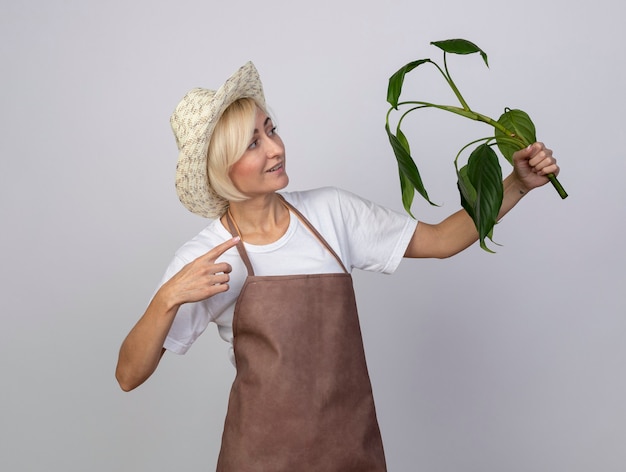 Femme souriante de jardinier blonde d'âge moyen en uniforme portant un chapeau tenant du doigt et regardant une plante isolée sur un mur blanc